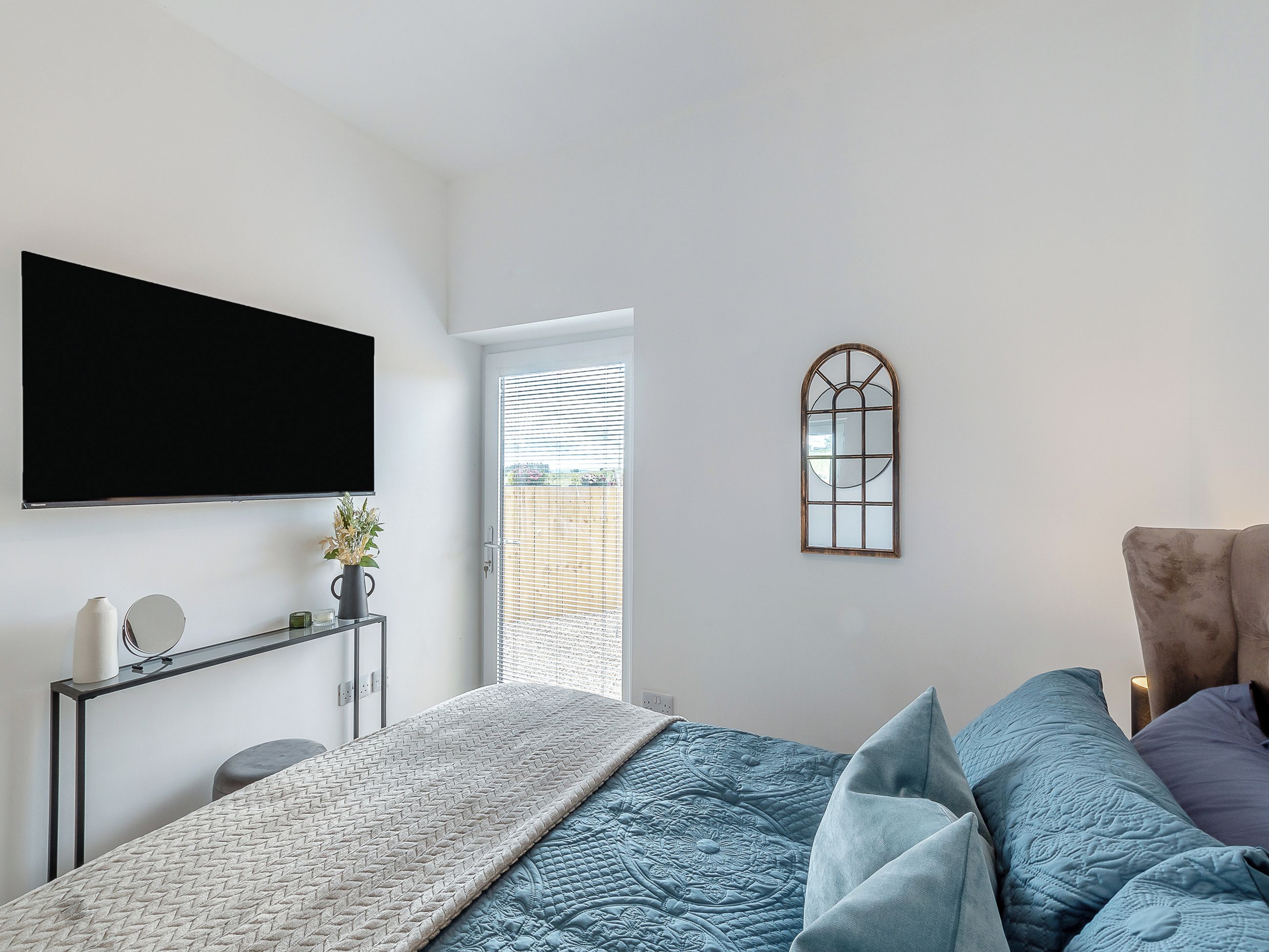 Une chambre à coucher comprenant un lit, un téléviseur fixé au mur, une table de chevet avec une lampe et une fenêtre avec des stores.