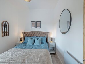 Un dormitorio con cabecera capitonada, cama con ropa de cama azul y blanca, dos mesitas de noche con lámparas, un espejo montado en la pared y dos cuadros enmarcados.