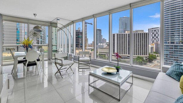 A modern living room with a glass coffee table, white sofas, and chairs, large windows with a city view, and a vase with flowers.