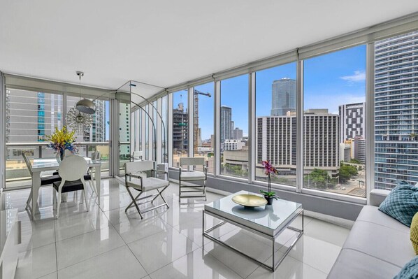 Una sala moderna con una mesa de centro de vidrio, sofás y sillas blancos, ventanas amplias con vista a la ciudad y un jarrón con flores.