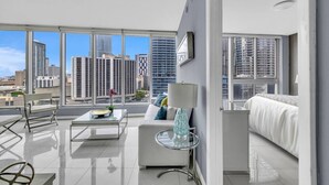 A modern living room with a sofa, coffee table, and a glass table with a lamp. Large windows offer a city view.
