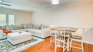 Uma sala de estar com sofá em formato de L, uma mesa de jantar com cadeiras, um pequeno banquinho e um tapete estampado.