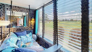 A bedroom with a stone-patterned wall, a bed with patterned bedding, a hanging chair, and a view of a grassy field through giraffe-patterned blinds. - Golfer’s Lava Cove - Waikoloa, HI (Waikoloa Village)
