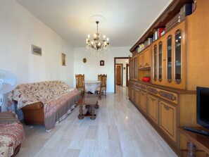 A living room with a sofa, a dining table with chairs, a chandelier, and a wooden cabinet with a television.