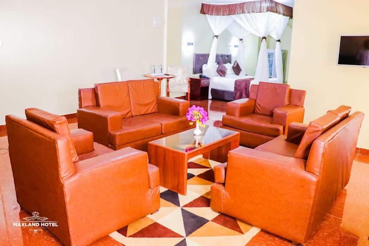 A hotel lobby with leather sofas arranged around a central coffee table, a bed with a canopy in the background, and a TV mounted on the wall.