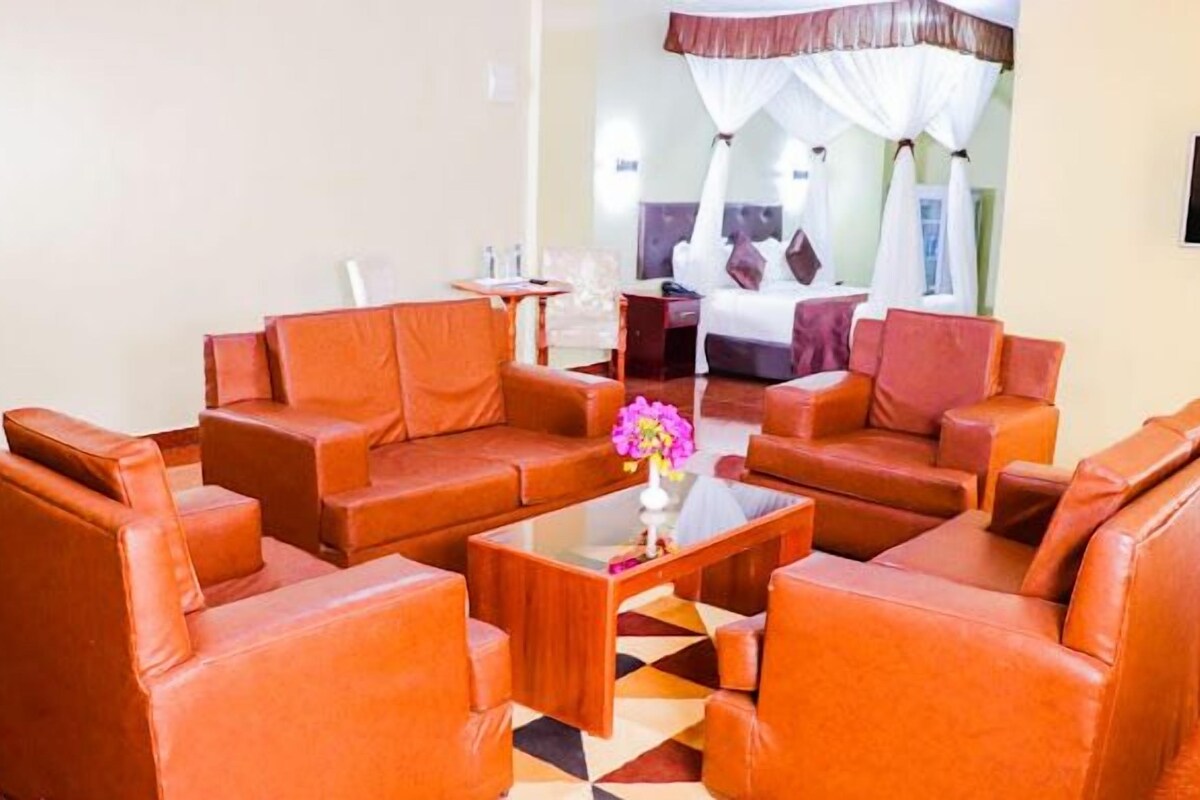 A hotel lobby with leather sofas arranged around a central coffee table, a bed with a canopy in the background, and a TV mounted on the wall.