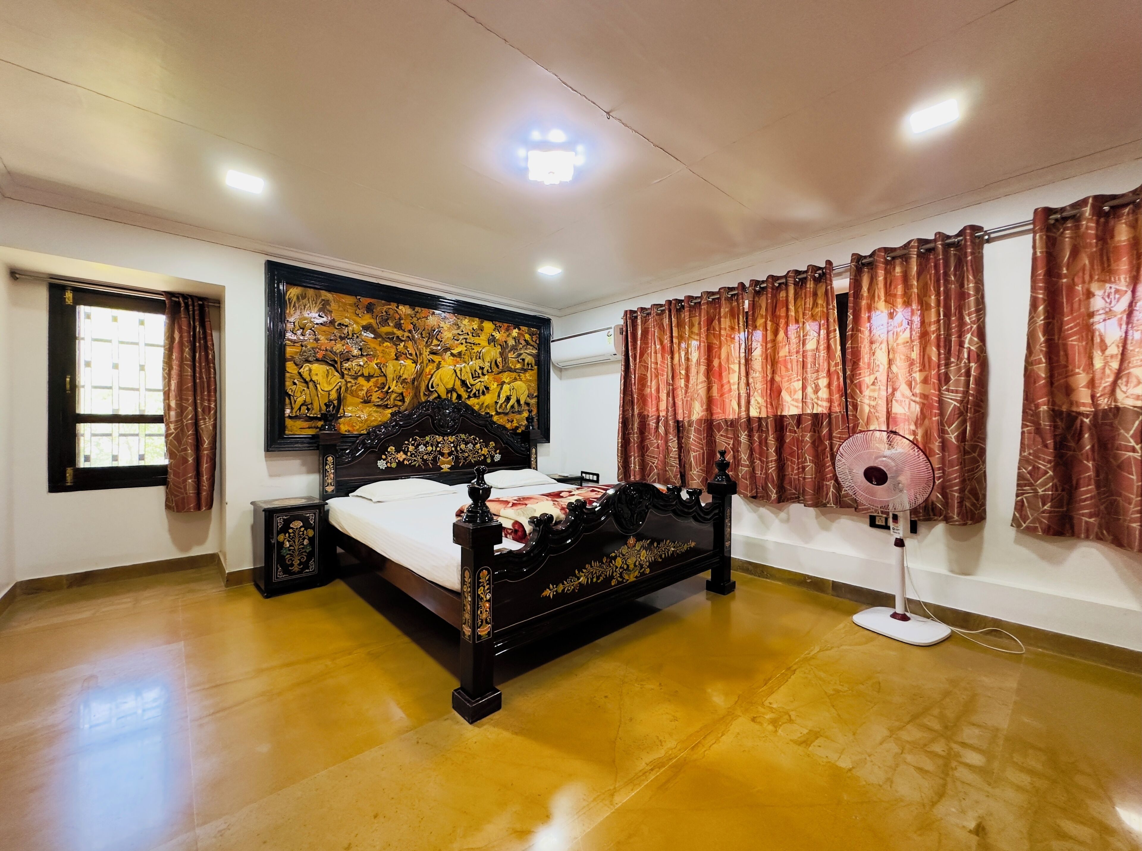A bedroom with a bed, a wall-mounted air conditioner, a standing fan, and a window with curtains.