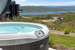 Uma banheira de hidromassagem com painel de controle, rodeada por um jardim exuberante e uma vista panorâmica de um lago e colinas.