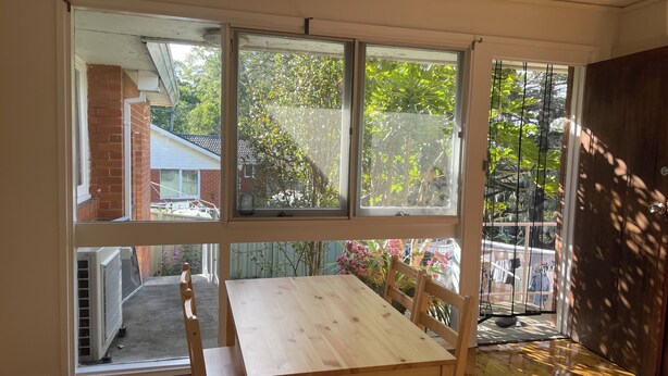 Une pièce avec une table et des chaises en bois, de grandes fenêtres et une vue sur un jardin et les maisons voisines.