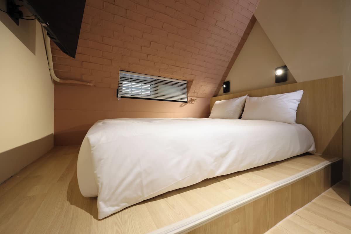 Un dormitorio con techo inclinado, cabecera de madera, cama blanca y ventana con persianas.