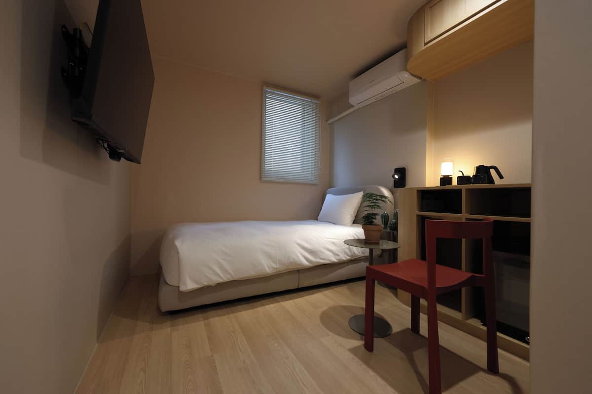 Un dormitorio pequeño y ordenado con una cama individual, un escritorio con silla, una ventana con persianas y un equipo de aire acondicionado.