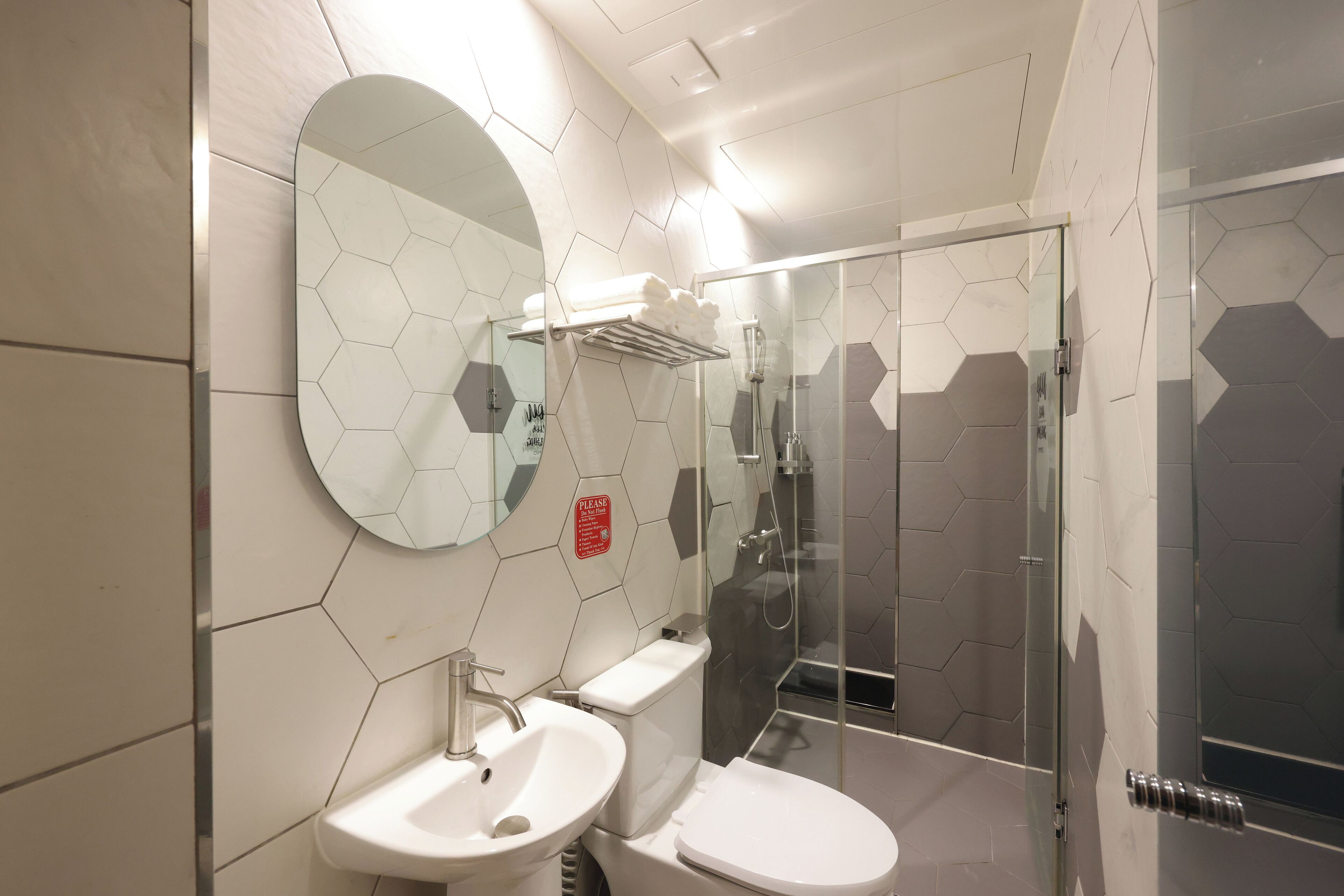 Un baño moderno con un patrón de baldosas hexagonales, un espejo redondo, inodoro, lavamanos y zona de ducha con mampara de vidrio.