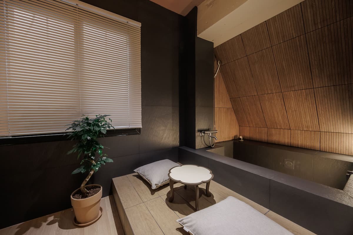 Un baño moderno con suelo de madera, una bañera grande, una planta en maceta y una mesita redonda con un cojín.