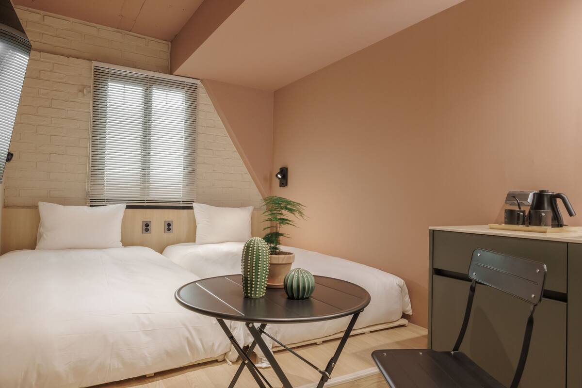Un dormitorio con cama, una mesa redonda, una silla, una planta en maceta y una cafetera.