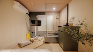Quarto de hotel compacto com cama, uma mesa pequena, televisão, chuveiro e uma planta em vaso.