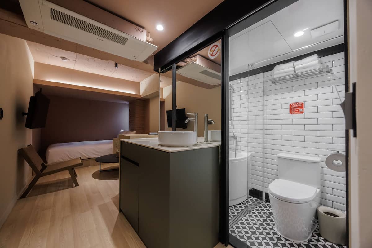 Una habitación de hotel compacta con cama, un pequeño rincón de descanso, kitchenette y baño con inodoro y lavamanos.