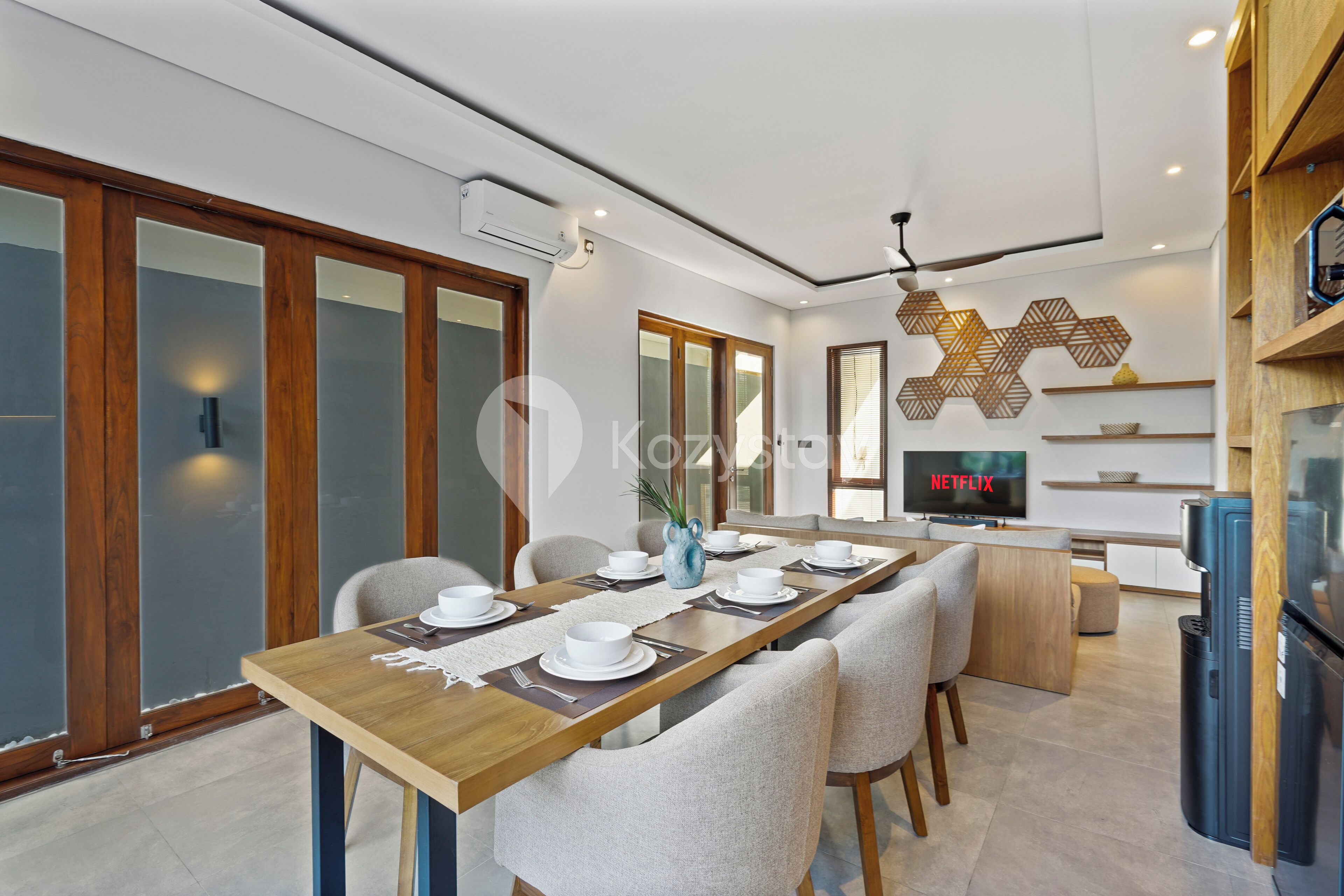 Un comedor moderno con una larga mesa de madera dispuesta para cuatro, sillas blancas y una cocina con un refrigerador y un televisor mostrando 'NETFLIX'.