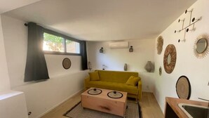A compact living space with a yellow sofa, a wooden chest, and a small kitchen area with a sink. - Cocoon under the oaks with jacuzzi (Saint-Martin-de-Londres)