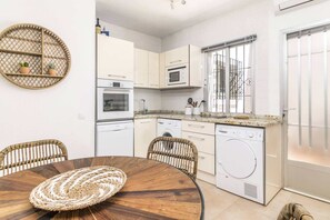 A compact kitchen with white cabinets, a microwave, oven, and a washing machine. There is a round dining table with a woven placemat.