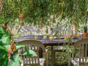 Een houten tafel met stoelen, omringd door groen, met glazen sinaasappelsap en fruit.