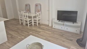 Ein modernes Wohnzimmer mit einem Fernseher auf einem weißen Schrank, einem Esstisch mit Stühlen und einem gewebten Korb auf einem weißen Tisch.