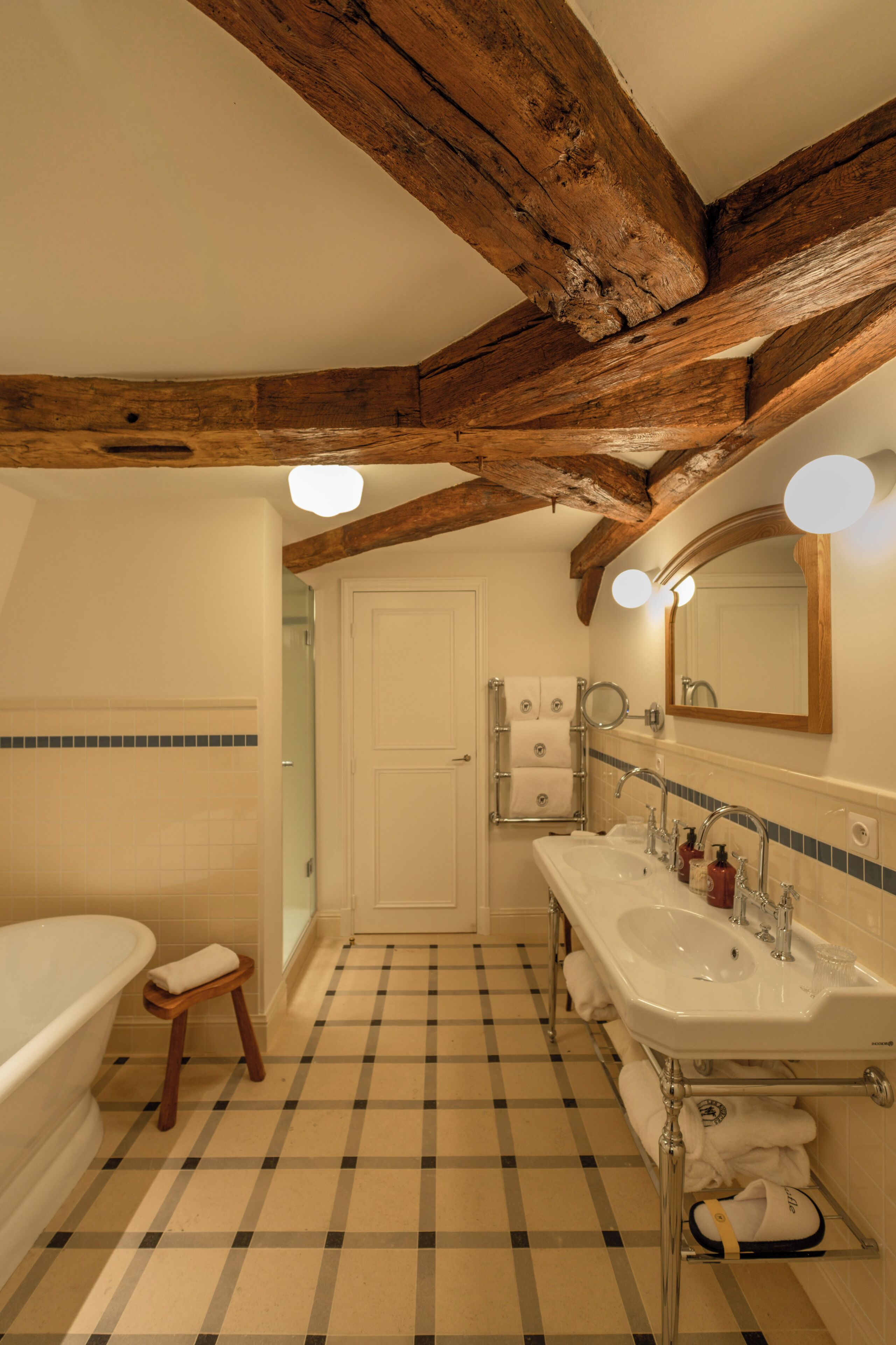 A bathroom with a checkered floor, wooden beams, a bathtub, a double sink, and a mirror with lights.