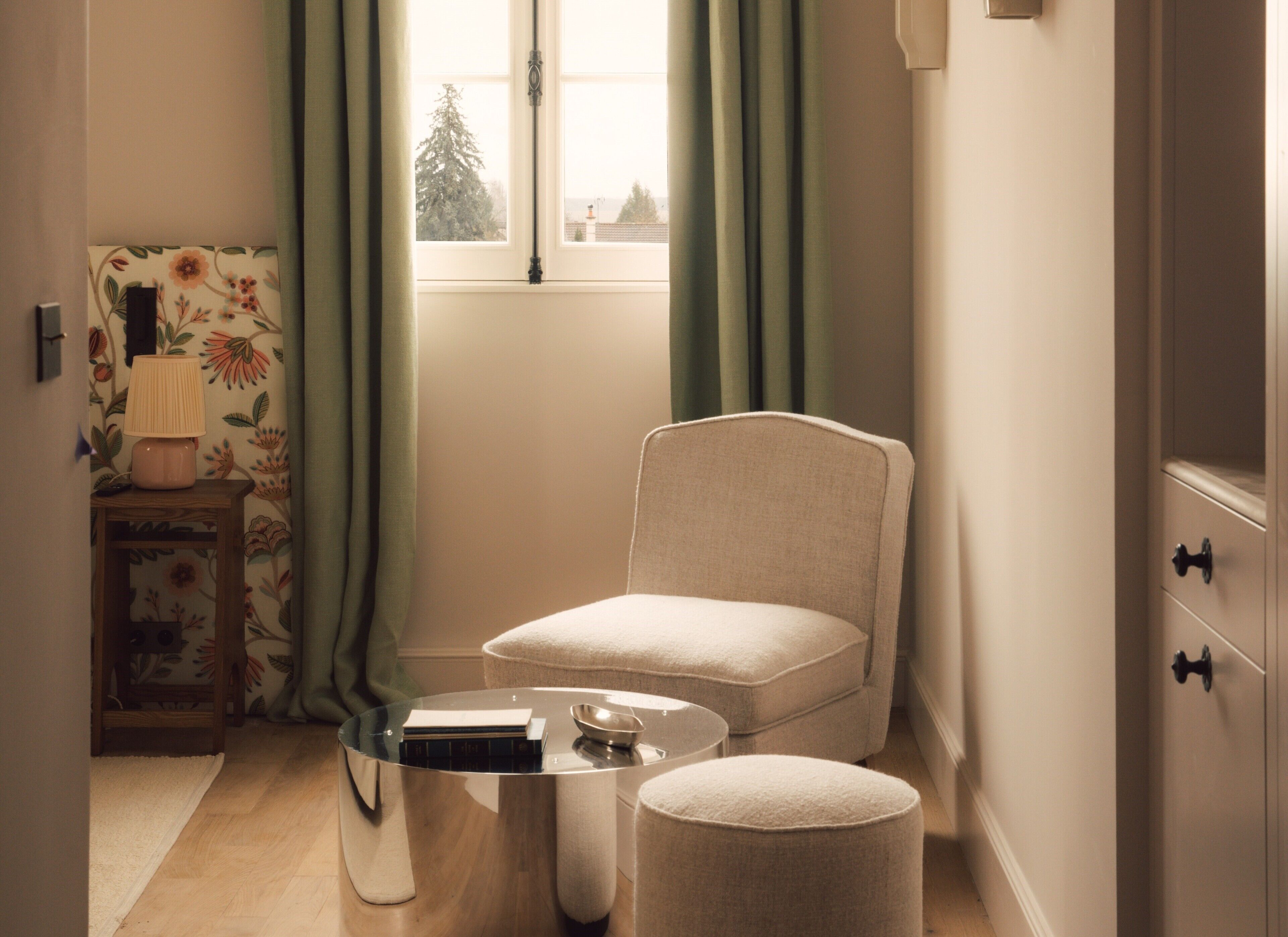 Ein gemütliches Zimmer mit Sessel, Hocker und einem kleinen Tisch mit Lampe.