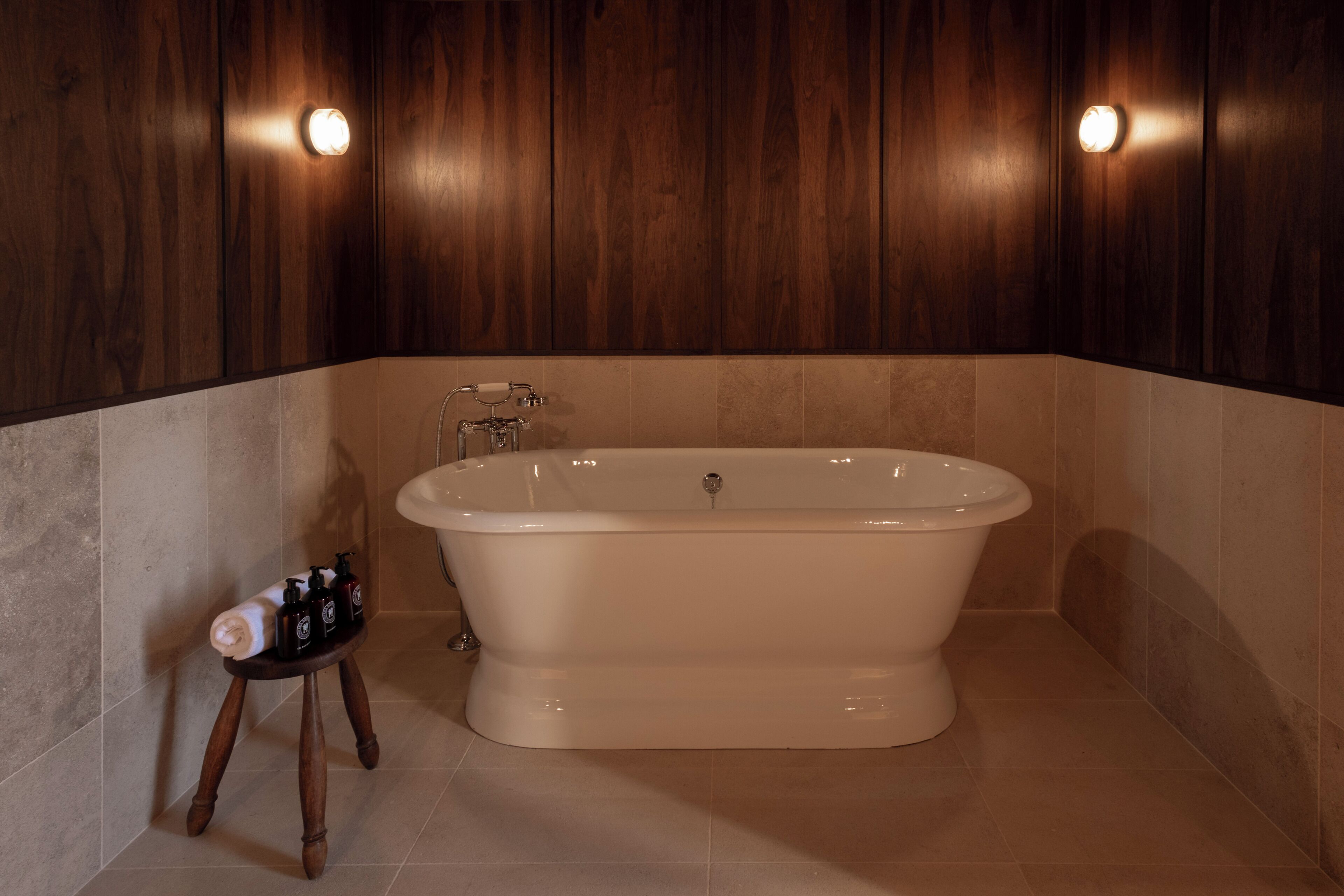 Een badkamer met een vrijstaand bad, houten wandbekleding en een klein krukje met toiletartikelen.