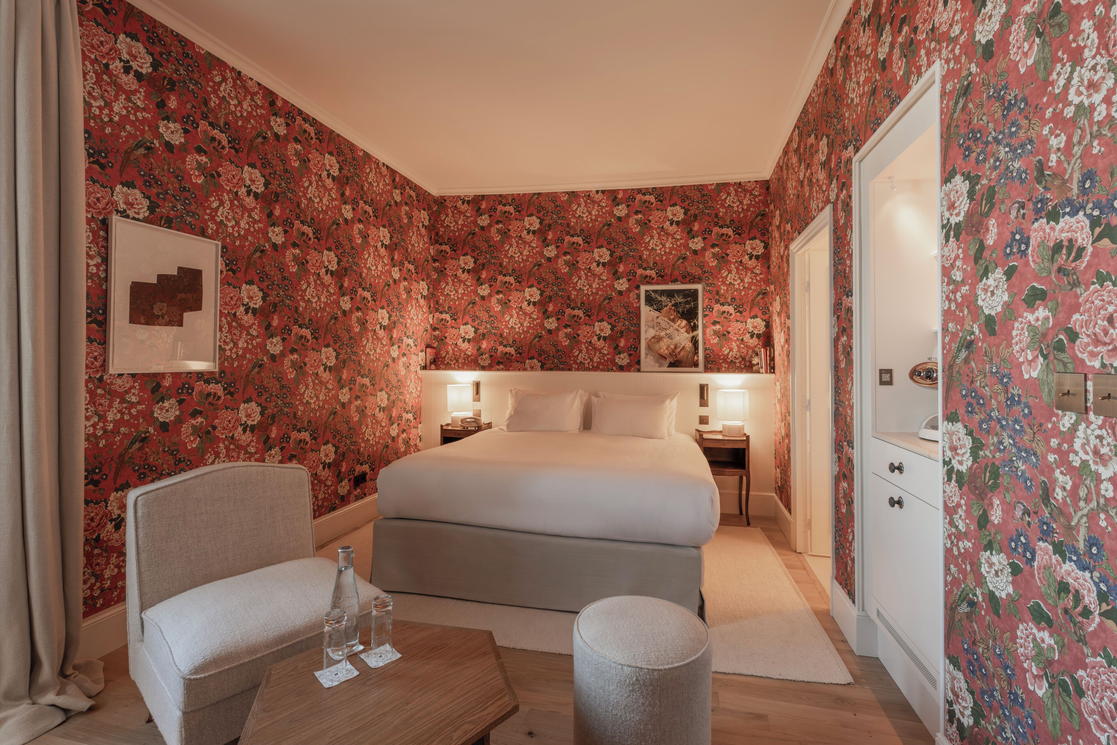 A bedroom with floral wallpaper, a bed, bedside tables, a chair, and a small table with a bottle and glass.