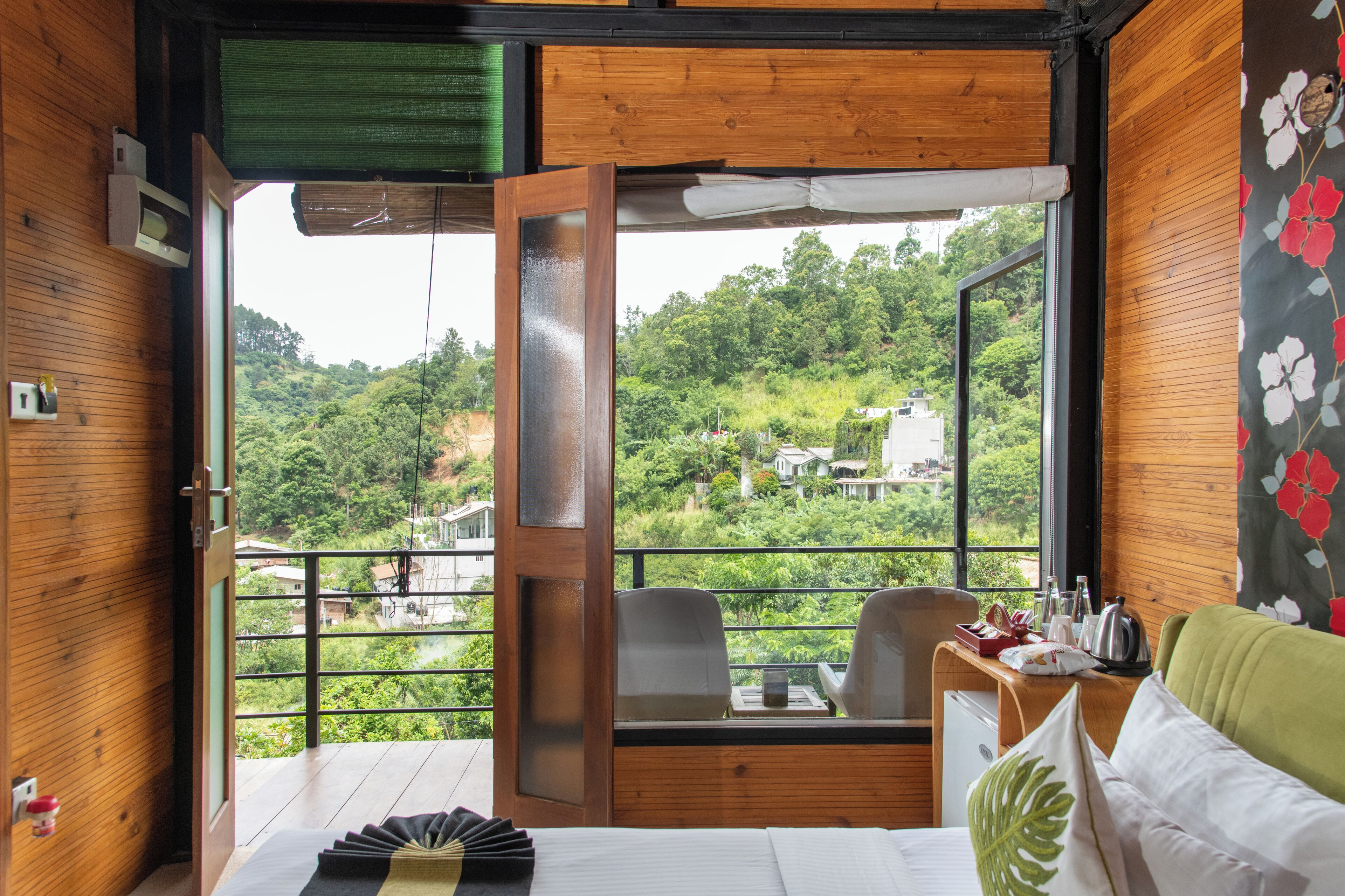 A room with a wooden interior, a balcony with a view of a green hillside, a bed with pillows, a desk with a chair, and a small table with a kettle and cups.