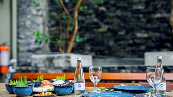 Una mesa de comedor servida con platos, tazones y vasos azules, con un muro de piedra y follaje al fondo.