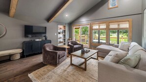 A modern living room with a large sectional sofa, two armchairs, a coffee table, and a dark entertainment unit. There is a ceiling fan, a large window with blinds, and a view of the outdoors.