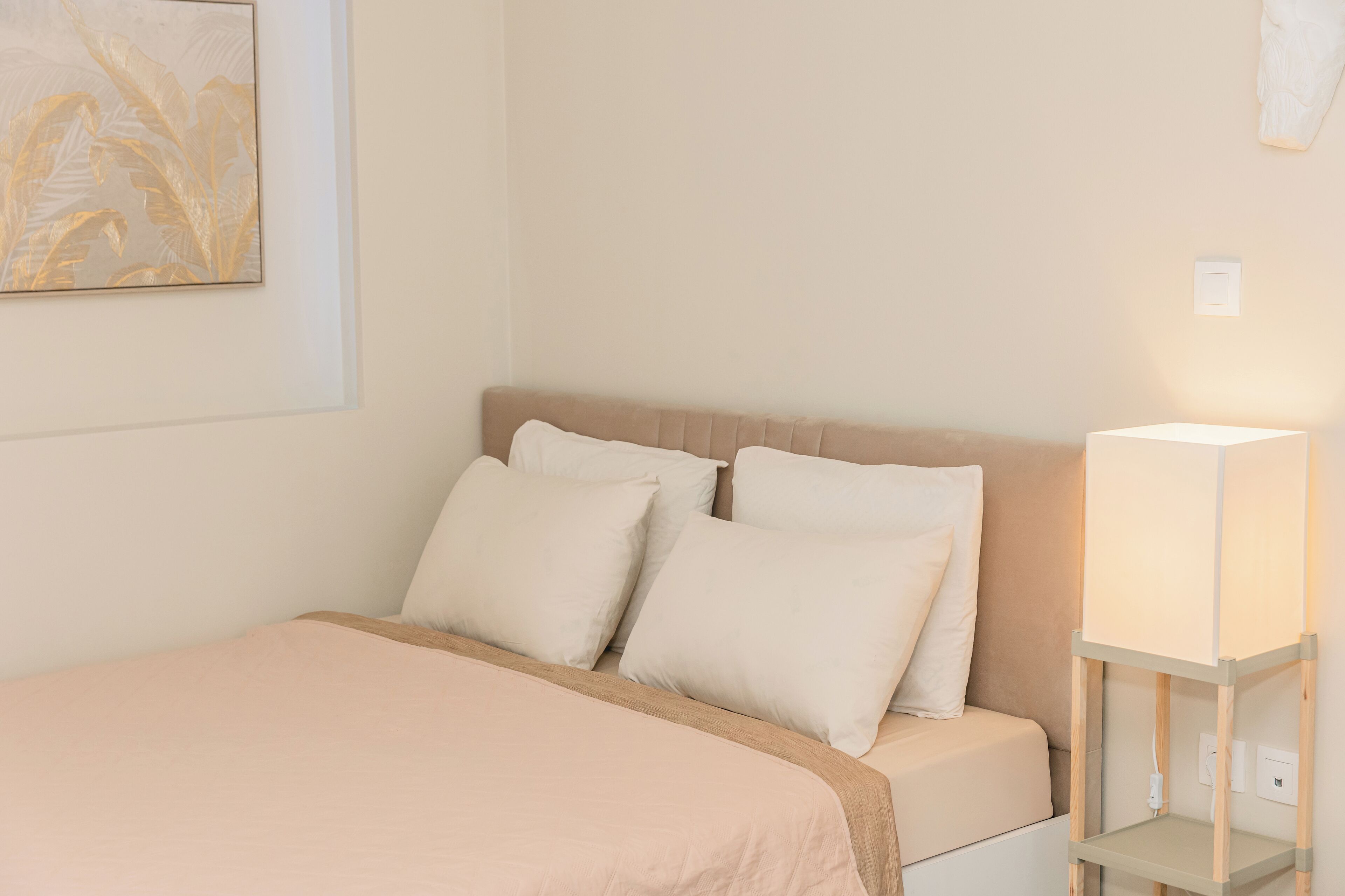 A neatly made bed with a beige bedspread and pillows, a bedside table with a lamp, and a wall-mounted light switch.
