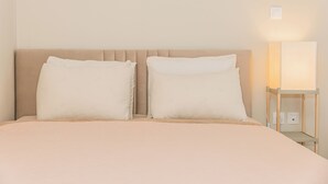 A neatly made bed with a beige headboard and two pillows, a bedside table with a lamp, and a wall-mounted light fixture. - Plantaea Studio - Athens Escape  (Athens)