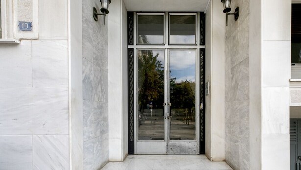 A building entrance with a marble floor, a glass door with a metal frame, and two wall-mounted light fixtures. - Plantaea Studio - Athens Escape  (Athens)