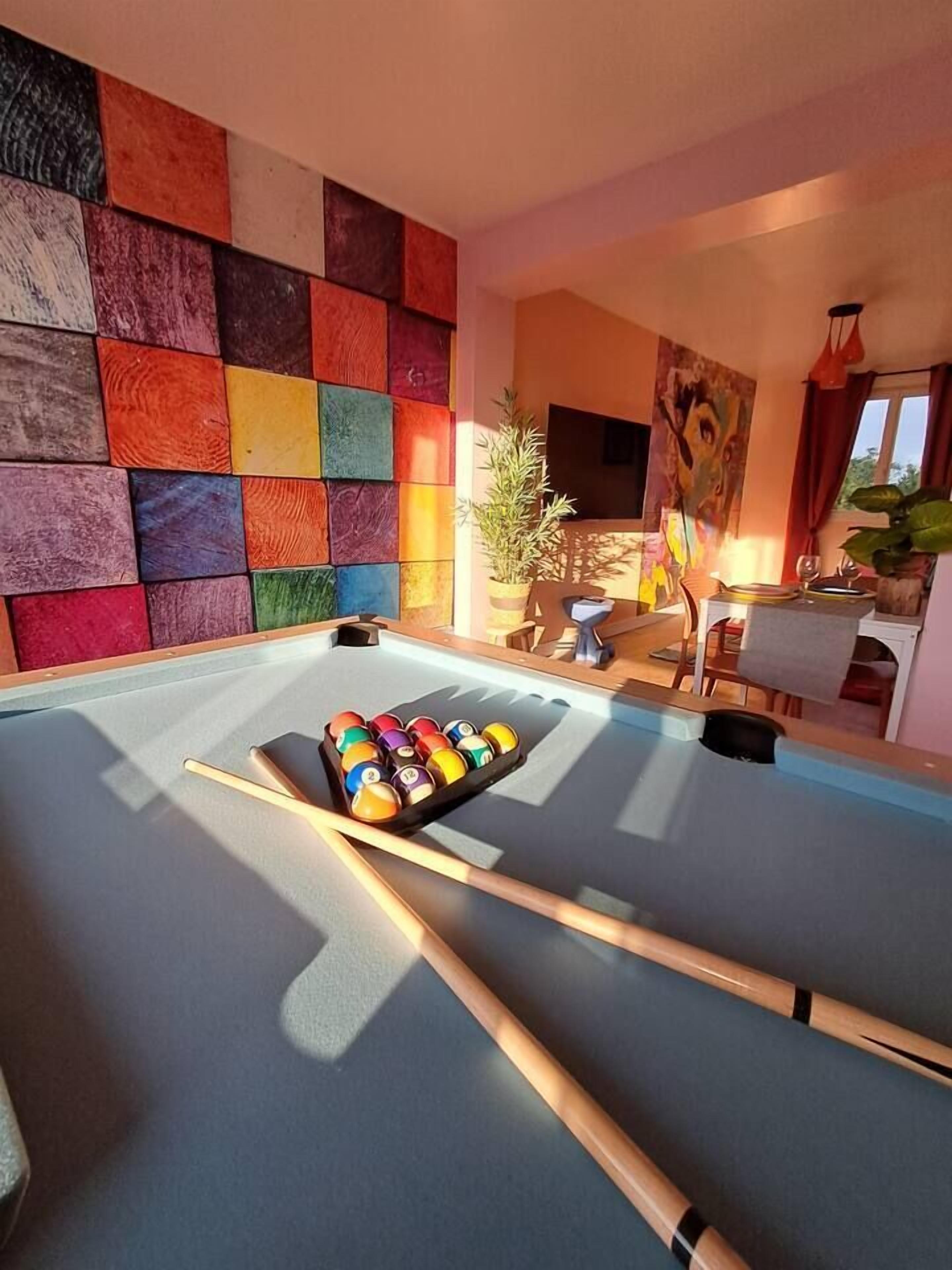 Ein Billardtisch mit Kugeln und Spielstangen, eine farbenfrohe Wand und ein Gemälde in einem Raum mit Essbereich.