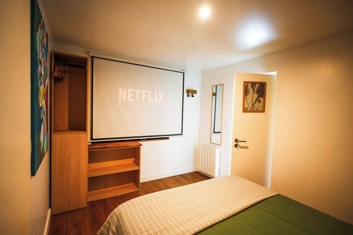 A bedroom with a large bed, a wooden wardrobe, a built-in TV screen displaying "NETFLIX", and a door with a mirror.