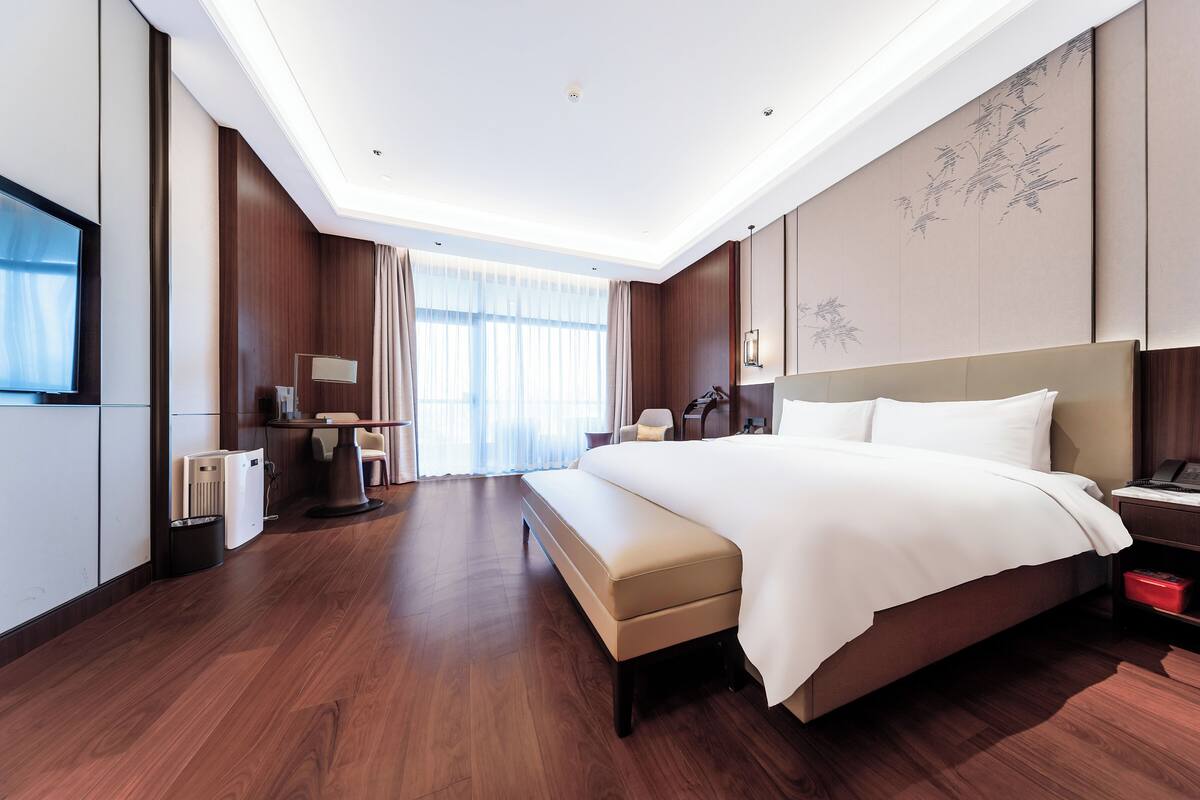A modern hotel room with a large bed, a desk with a lamp, a chair, and a television.
