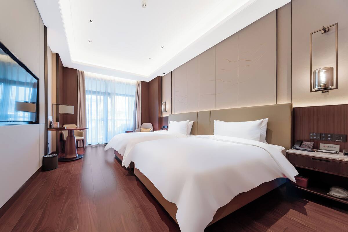 A hotel room with two beds, a desk with a telephone, and a large window with curtains.
