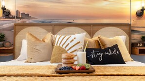 A bed with a cityscape view, a tray of fruit and donuts, and a pillow with 'stay awhile' text.