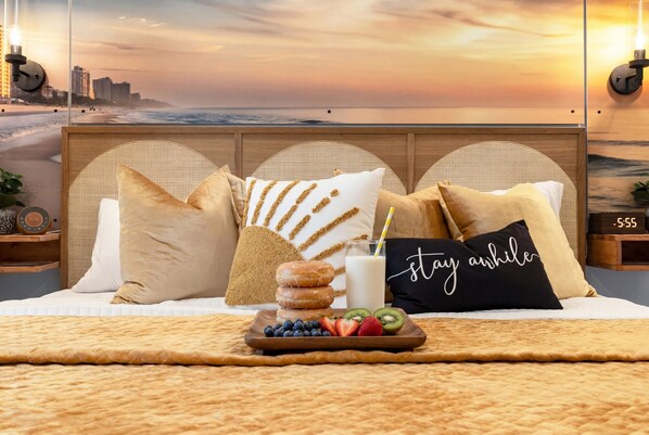 A bed with a cityscape view, a tray of fruit and donuts, and a pillow with 'stay awhile' text.
