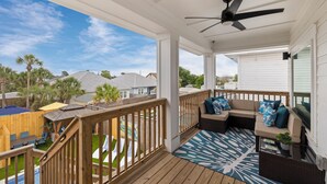 A covered outdoor deck with a ceiling fan, a sofa with cushions, and a view of a swimming pool and neighborhood houses.