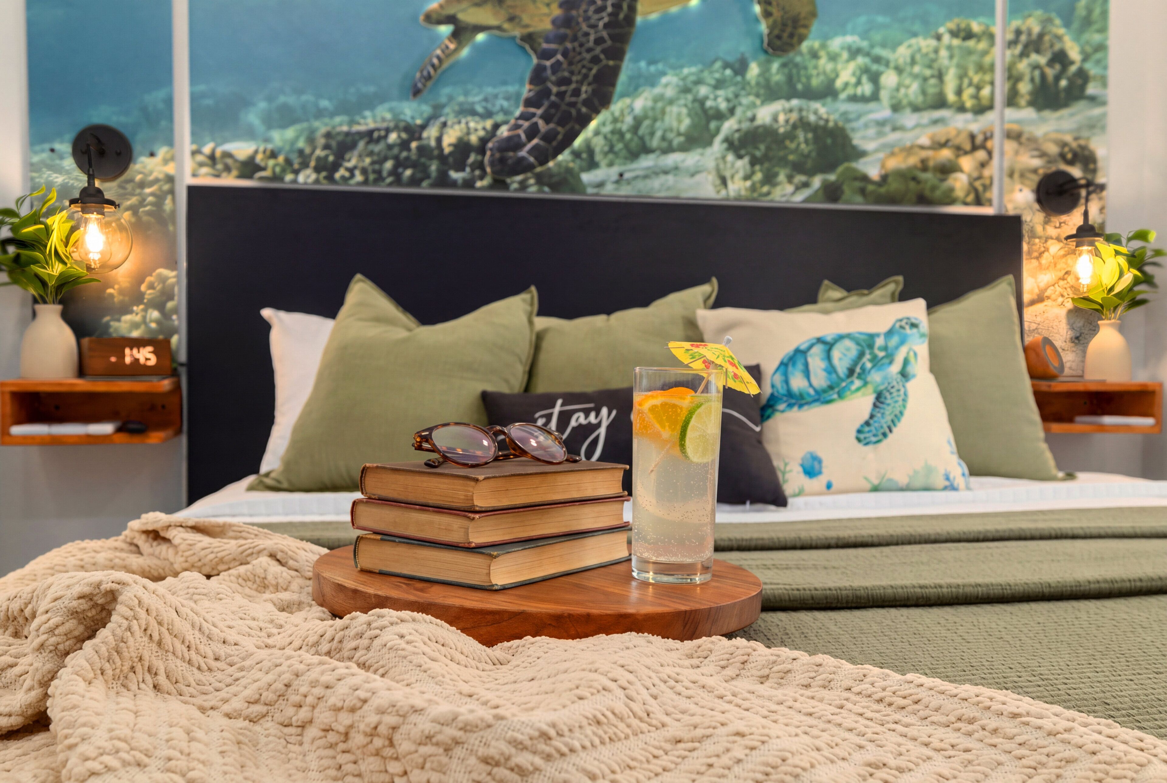 Una cama con cabecera, almohadas, una mesita de noche con un reloj, libros y un vaso de bebida, además de un mural en la pared.