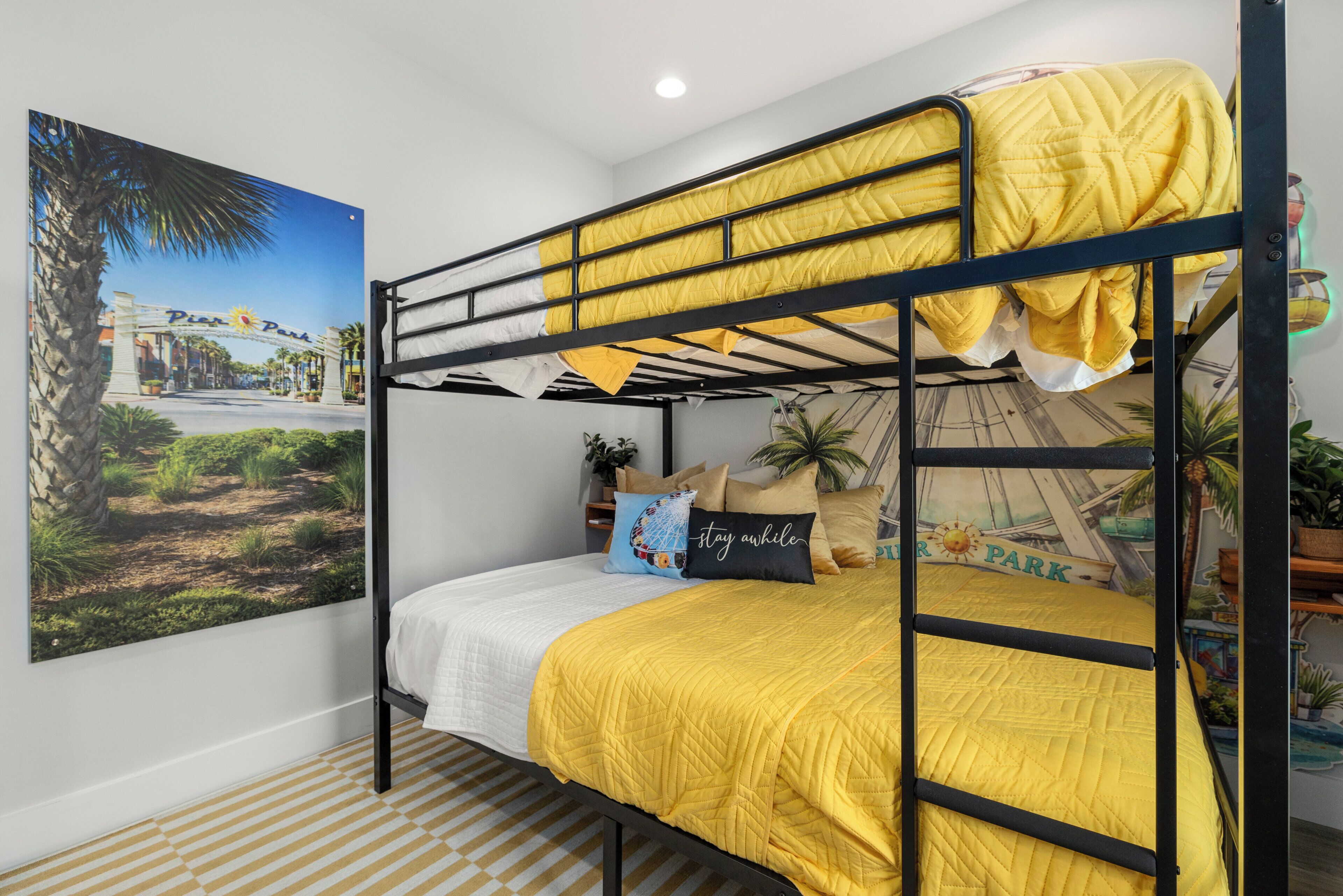 Un lit superposé avec une couette jaune, une couette blanche et un coussin sur lequel est brodé le mot « restez un peu ». Un poster représentant une rue ensoleillée est accroché au mur.
