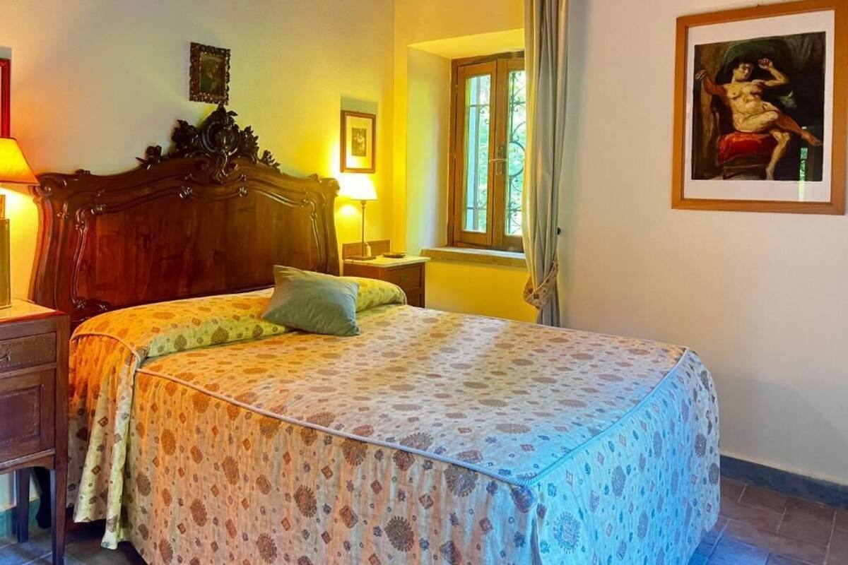 A bedroom with a wooden bed, a bedside table, a lamp, a window with curtains, and a framed picture on the wall.
