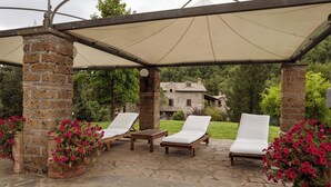 A covered outdoor seating area with stone flooring, two lounge chairs, a wooden table, and potted plants. - Borgo La Ferriera (Celleno)
