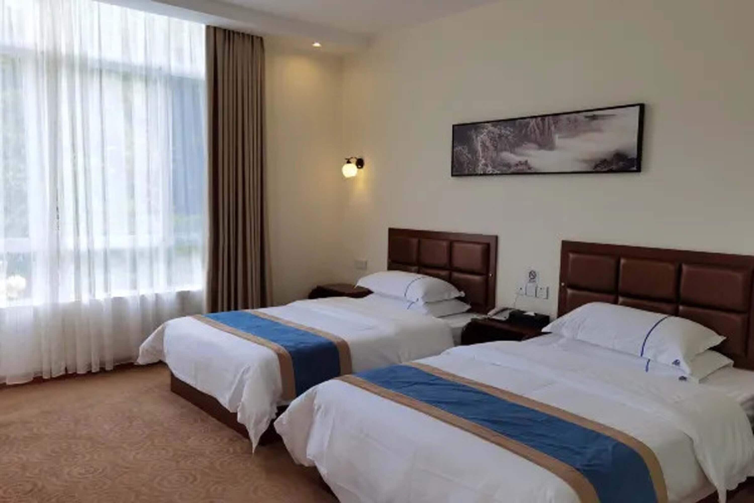 A hotel room with two beds, a large window with curtains, a wall-mounted light, and a painting.