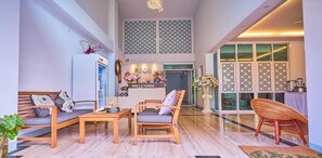 Un grand espace de vie comprenant un canapé, des fauteuils et une table basse. On aperçoit, au fond, un comptoir d'accueil avec une pancarte indiquant « BIENVENUE ».