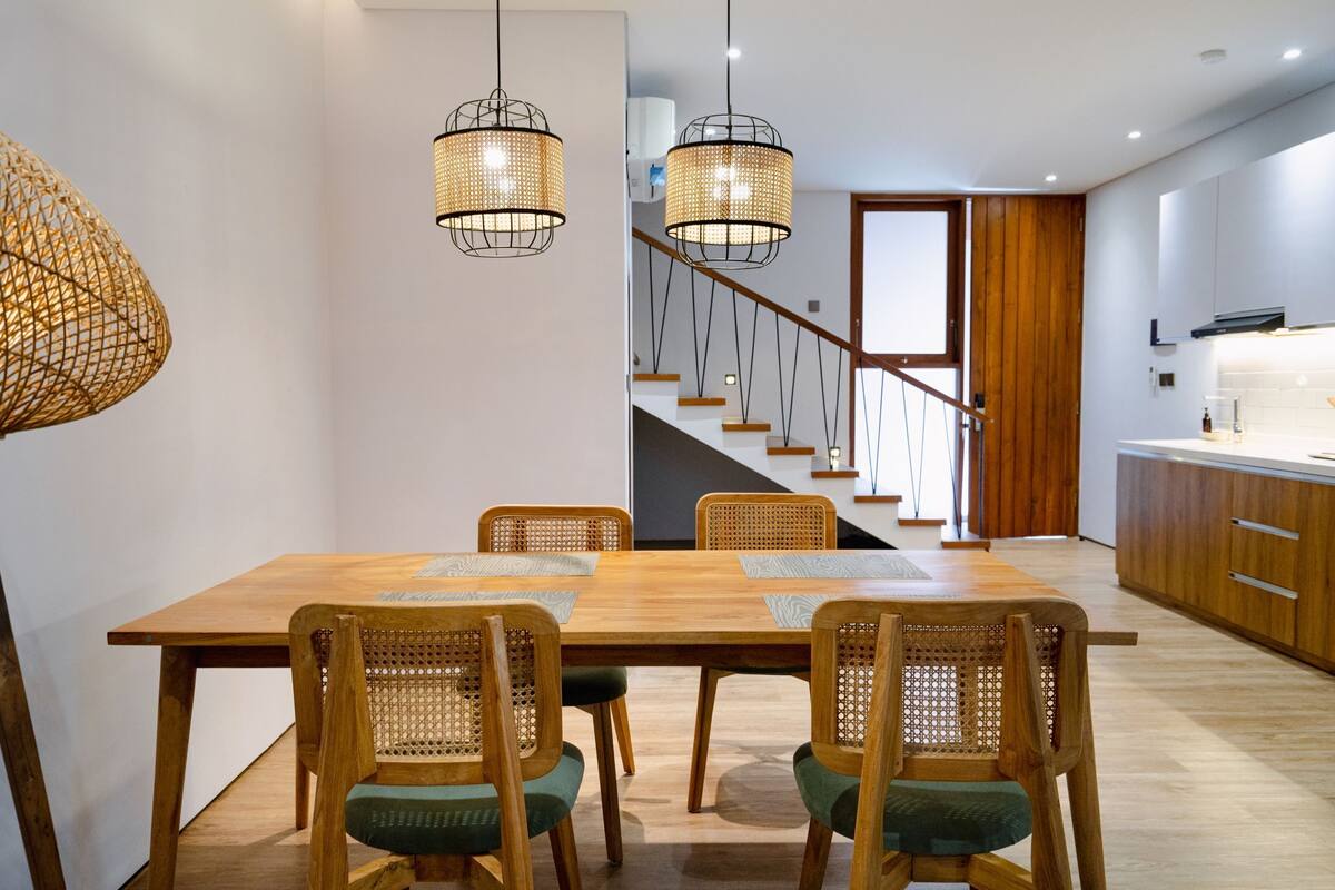 Un espace repas moderne avec une table et des chaises en bois, un escalier en arrière-plan et un coin cuisine doté de meubles en bois.