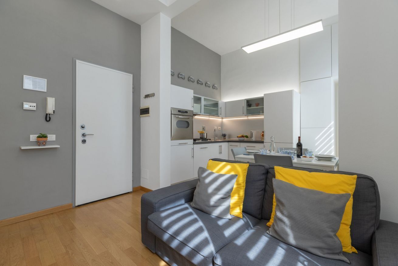 Een moderne woonkamer met een grijze bank, een keuken op de achtergrond en een witte deur.
